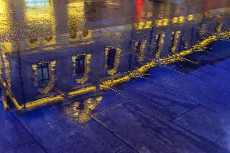 Water on stone sidewalk reflects a purple building rimmed in yellow light