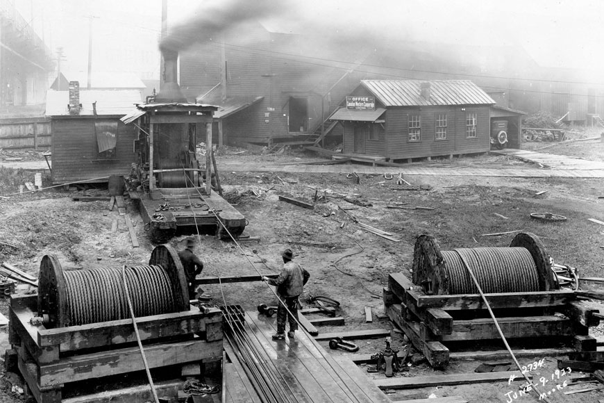 B&W photo shows steam engine almost 2 stories high with cables running from and workers looking on