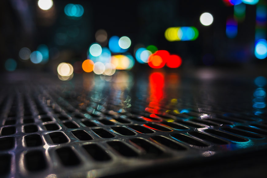 A closeup shot of a wet storm drain grate at night, black but reflecting lights in the background