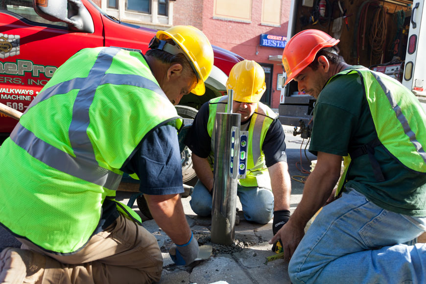 The installation crew finishes filling a hole, holding a steel pipe bollard, with concrete.