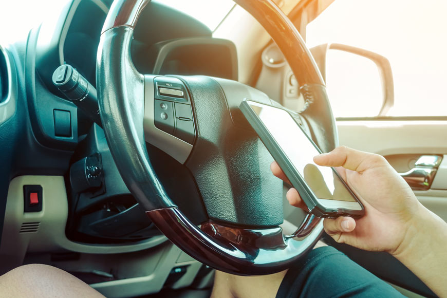 A close shot of a hand holding a phone and texting near a steering wheel.