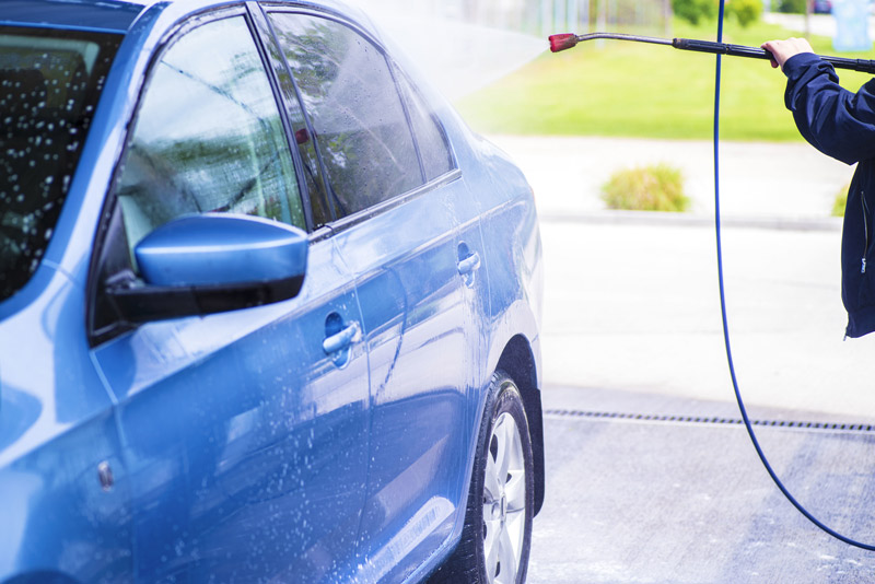 A person pressure washes a blue car in a driveway