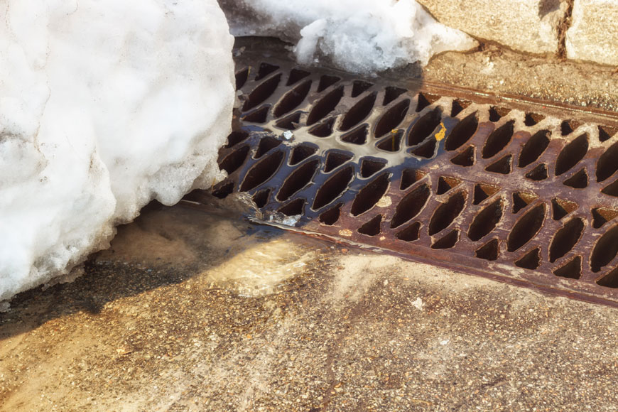 A brown cast iron trench grate sits beside a curb under a lump of snow