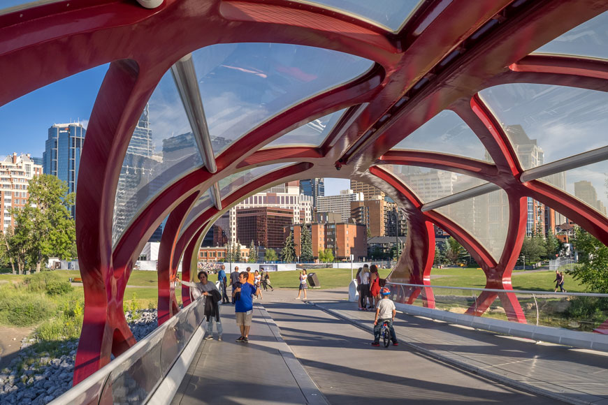 A tube-like bridge is made of red steel girders that wrap in an argyle pattern around pedestrians and cyclists