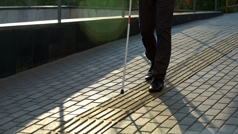 A man with a cane uses wayfinding tenji blocks to make his way down a city street at sunset